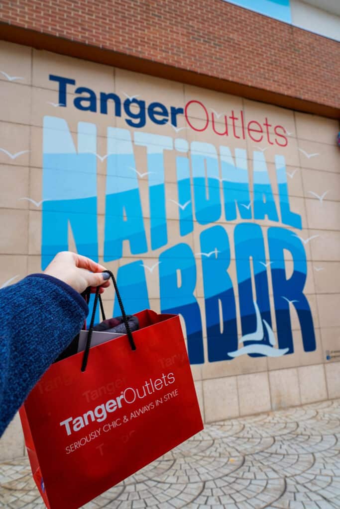 A woman's hand holding a red Tanger Outlets shopping bag in front of a Tanger Outlets National Harbor mural