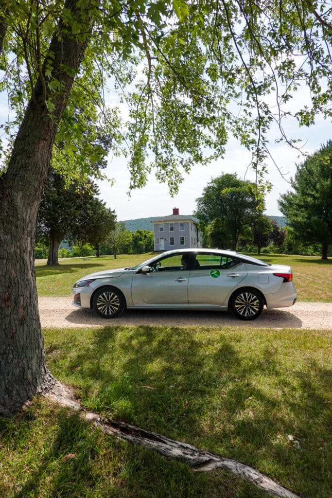 Scenic photo of a silver Zipcar sedan parked under a tree on a dirt road